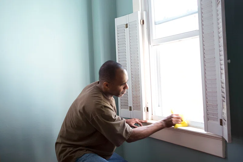 A man cleaning a window sill with a yellow cloth inside a room.